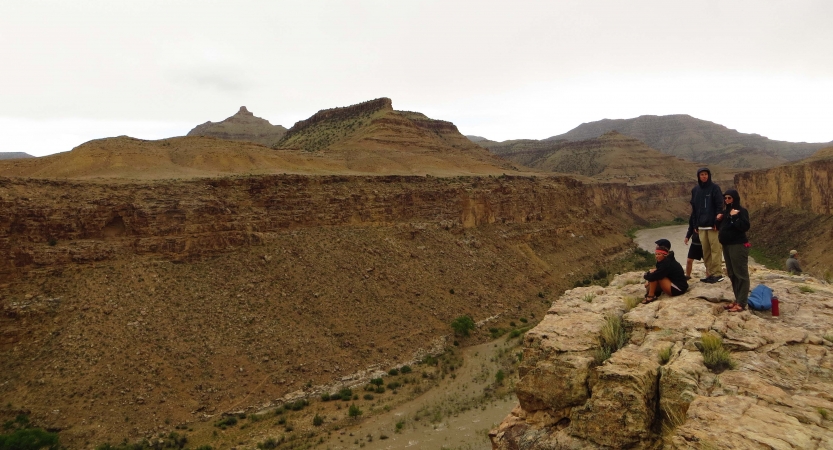 Three people are gathered on a cliff above a river between high canyon walls.